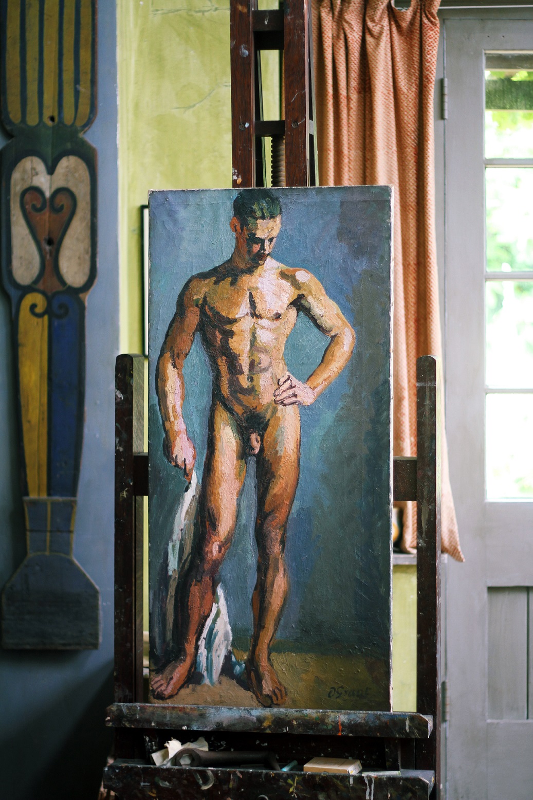Duncan Grants painting of a standing male nude rests on an easel in the studio at Charleston. You can see a curtain and a door in the background on the right, and a decorative work on the wall in the background on the left.