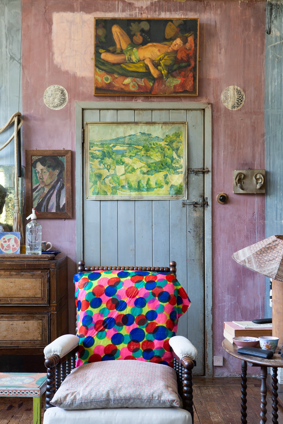 A photograph of the studio at Charleston. A chair covered in a colourful fabric sits in front of a wooden door painted a pale blue. Above the door is a painting of a male in green pants reclining by Duncan Grant.