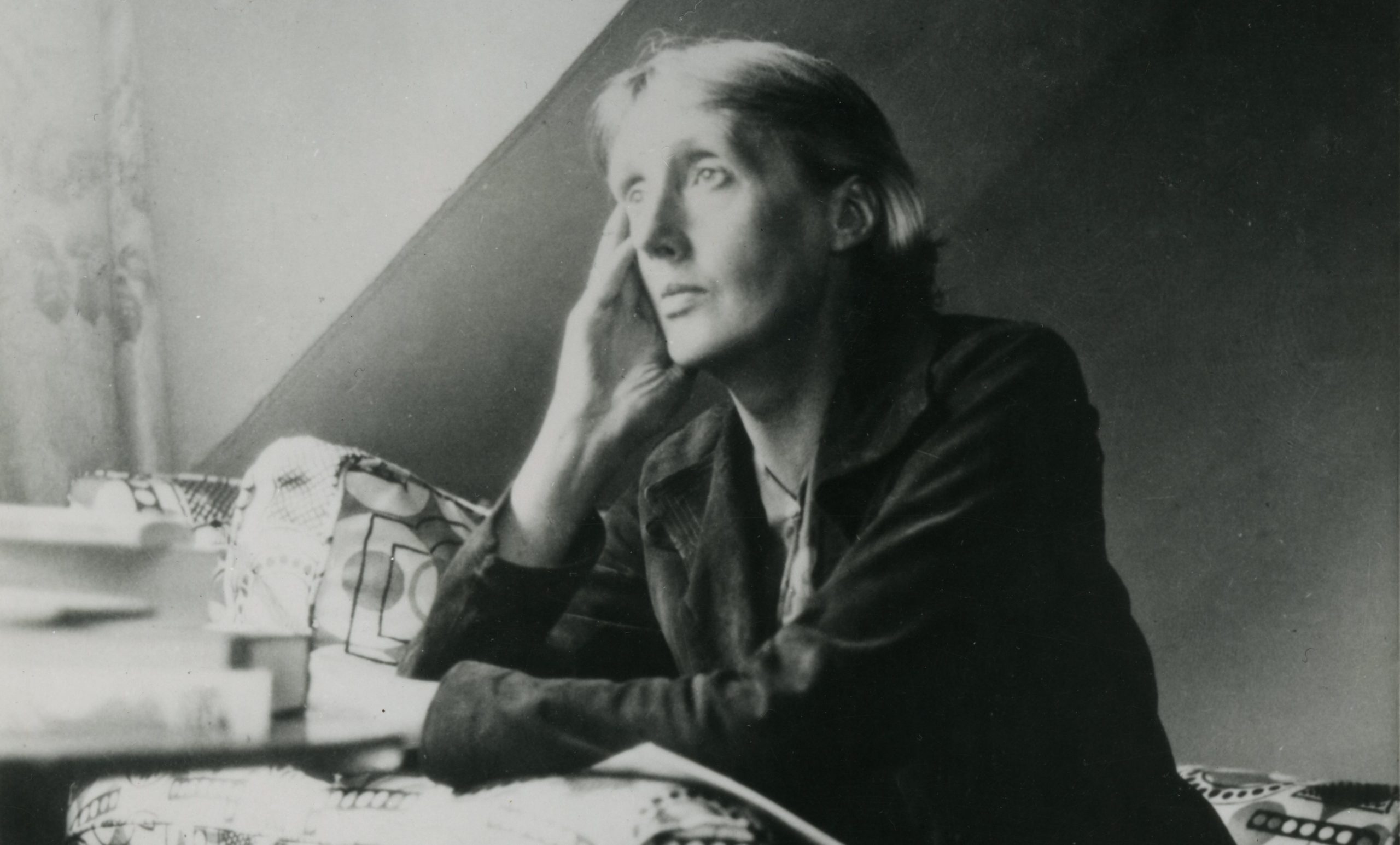 A black and white photograph of Virginia Woolf looking out of a window whilst sitting in an armchair upholstered in patterned fabric. She is resting her face on her right hand as she looks. Her other arm is folded across in front of her. She has he short hairstyle tucked behind her ears.