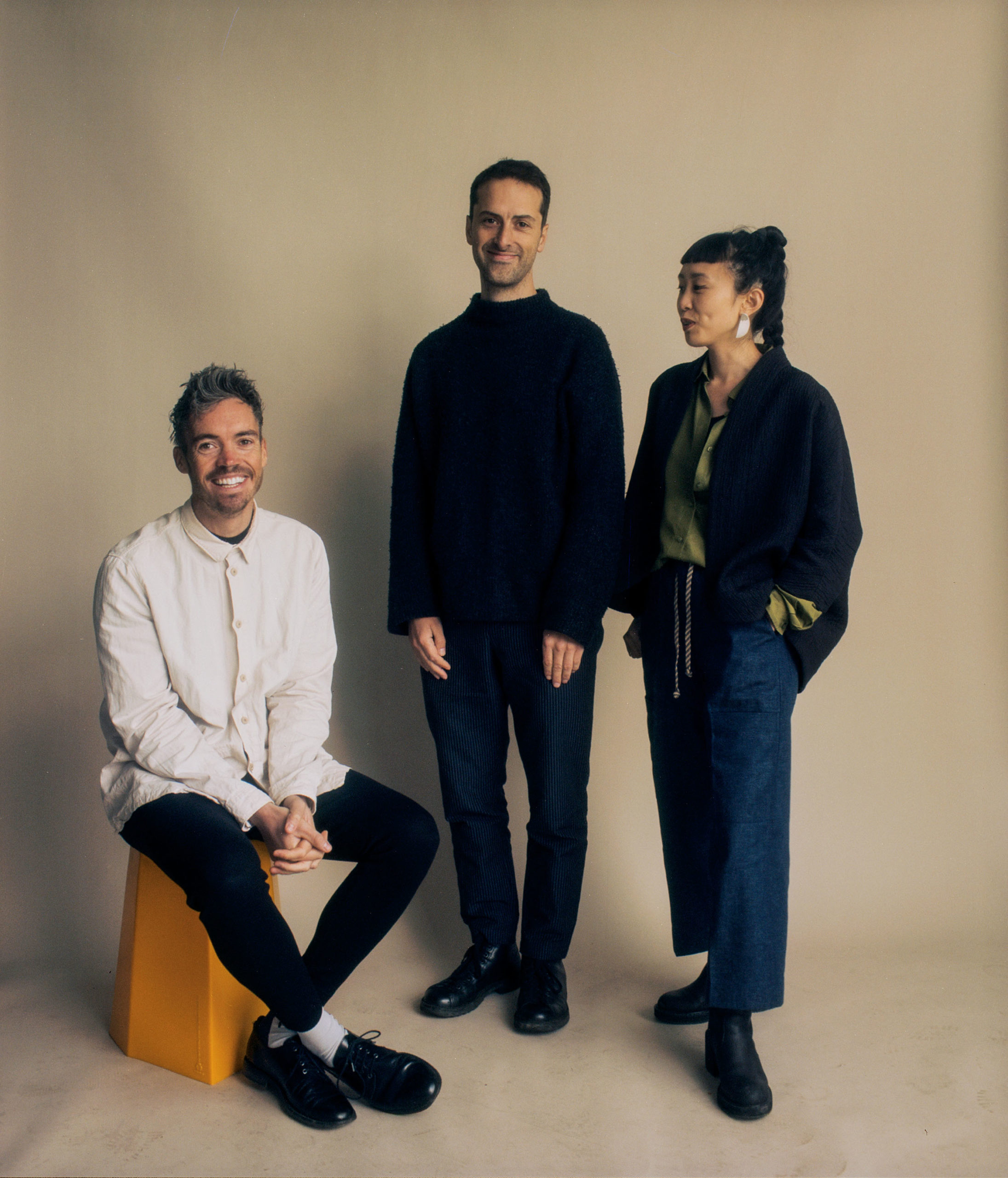 A portrait photograph of three people. The man on the left in a white shirt sits on a plinth with his legs crossed. The man in the middle is all in black. The woman on the right is looking to the left of the frame and has her hands in her pockets.